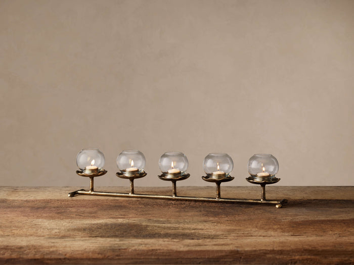 Artemis Multi Candle Holder. Horizontal antique brass stand holds 5 lit tealights in clear glass globes on a rustic wood table.