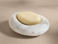 A white Carrara marble soap dish with smooth veining, holding an oval beige soap, placed on a neutral surface.