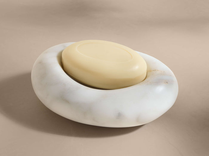 A white Carrara marble soap dish with smooth veining, holding an oval beige soap, placed on a neutral surface.