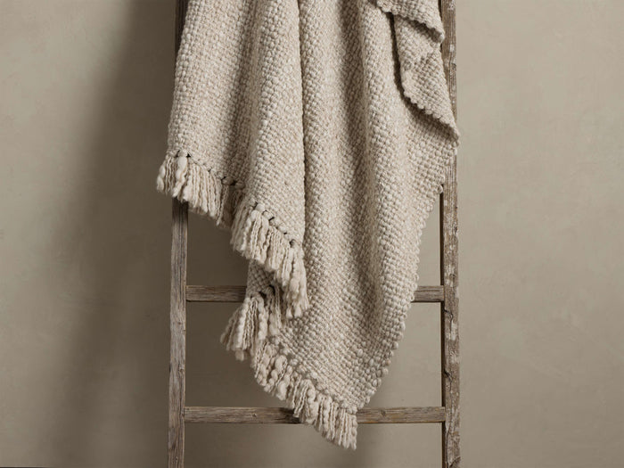 Light beige chunky knit throw blanket with bobble texture and fringed edges, casually draped over a rustic wooden ladder.