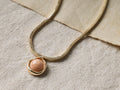 Close-up of the gold snake chain Aurette Necklace with a round peach-colored stone pendant in a gold bezel.
