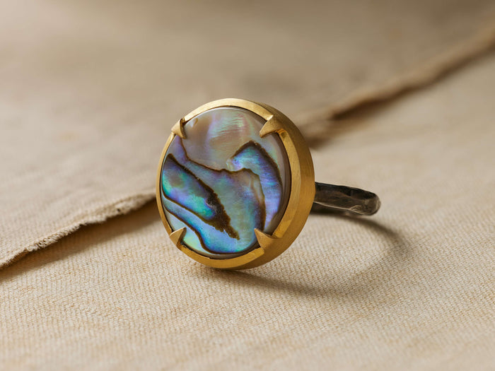 Autry Abalone Ring with iridescent oval abalone stone in 14k gold setting on oxidized silver hammered band, on light fabric.