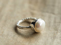 A sterling silver ring featuring a large, round freshwater pearl on a textured band, resting on a beige fabric surface.