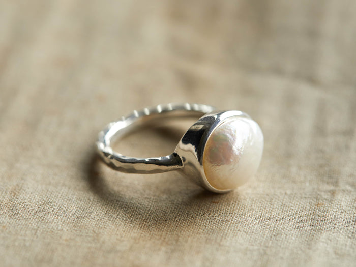 A sterling silver ring featuring a large, round freshwater pearl on a textured band, resting on a beige fabric surface.