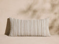 Avola Outdoor Lumbar Pillow, rectangular, with vertical white and light brown stripes, resting on a beige surface against a wall.