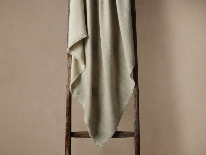 Light gray Baby Alpaca Throw in Fog, with fringed edges, draped over a rustic wooden ladder against a neutral wall.