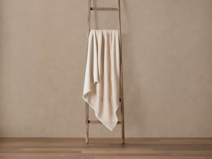 Light beige Baby Alpaca Throw with fringed edges draped over a rustic wooden ladder, against a warm beige wall and wood floor.