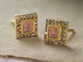 Close-up of two gold Beatrice Earrings. Each square earring features an outer border of brown diamonds surrounding a pink textured center.