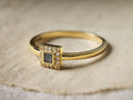 Close-up of a Gold Beatrice Ring with a square dark center stone surrounded by clear accent stones.