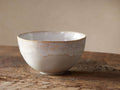 Single Bela Cereal Bowl, creamy off-white stoneware with reactive glaze and subtle golden-brown antiquing on a wood surface.