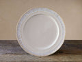 Creamy off-white Bela dinner plate with an antiqued reactive glaze rim, resting on a rustic wooden table.