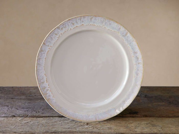 Creamy off-white Bela dinner plate with an antiqued reactive glaze rim, resting on a rustic wooden table.