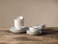 Bela Dinnerware set: Stacked creamy off-white, glazed stoneware bowls & plates, with mugs & saucers on a wooden table.