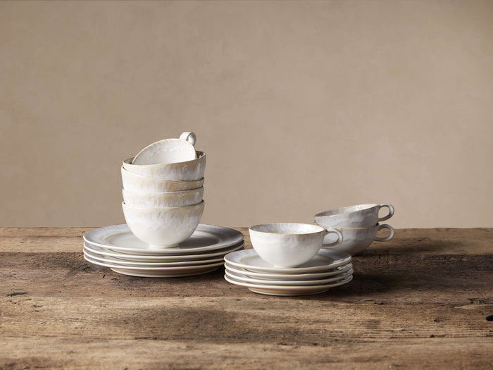 Bela Dinnerware set: Stacked creamy off-white, glazed stoneware bowls & plates, with mugs & saucers on a wooden table.