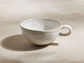 Bela stoneware mug, creamy off-white with reactive glaze showing subtle tan/pinkish marbling, handle on right.