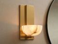 Belmira Sconce: Brass-finished wall light with glowing, bowl-shaped alabaster shade on a textured wall next to a mirror.
