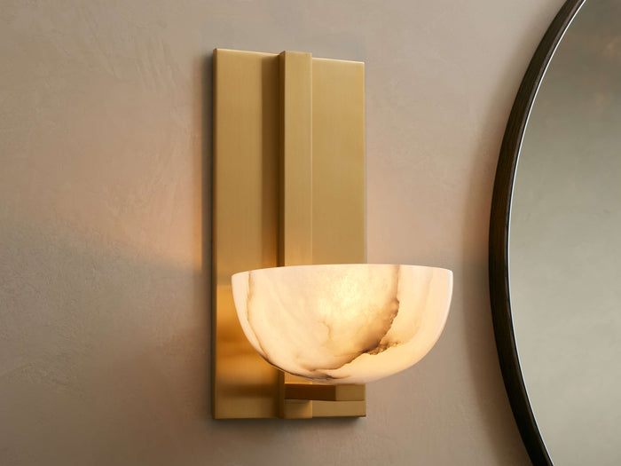 Belmira Sconce: Brass-finished wall light with glowing, bowl-shaped alabaster shade on a textured wall next to a mirror.