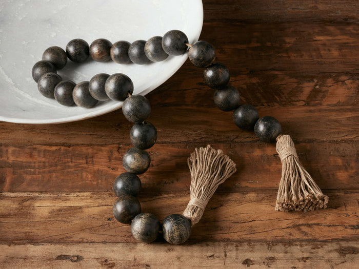 Black beaded strand made of distressed mango wood beads on jute rope, with tassels, resting on a wooden surface beside a white bowl.