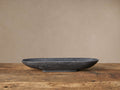 Black Marble Oval Bowl, elongated and dark grey, sits horizontally on a rustic wooden table against a light beige wall.