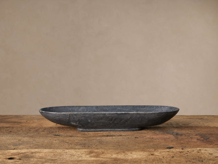 Black Marble Oval Bowl, elongated and dark grey, sits horizontally on a rustic wooden table against a light beige wall.