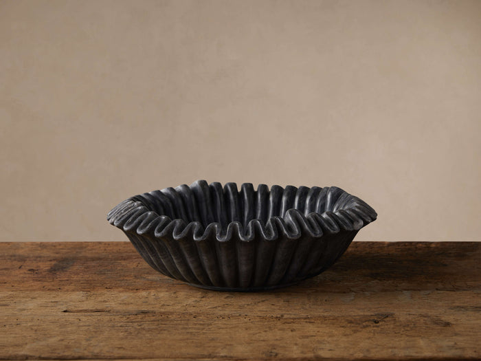 Black marble ruffle bowl with wavy edges, featuring natural grey tones, sits on a rustic wooden table against a light wall.