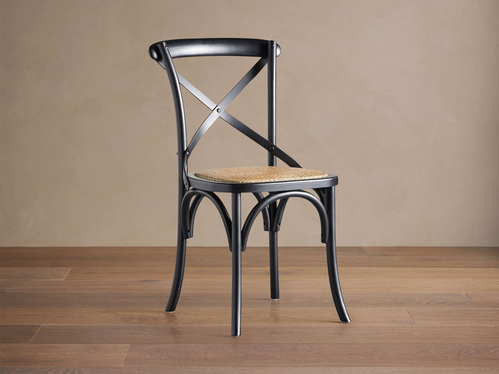 Black Cadence Dining Chair with natural rattan seat. X-back, curved legs, angled on a brown wood floor against a beige wall.