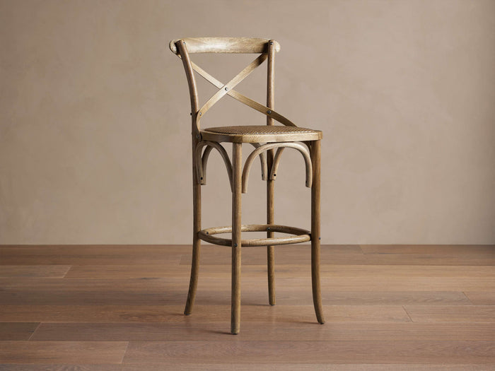 Cadence Stool, natural weathered oak with X-back and curved legs, light brown handwoven rattan seat, on wood floor.