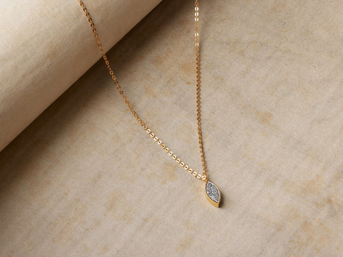 Cairn Elliptic Necklace with a gold chain and an elliptic pendant featuring raw diamonds, displayed on a textured beige surface.