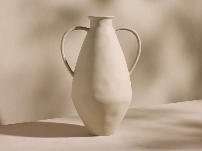 A matte white ceramic vase with a tall, slightly uneven silhouette and two curved handles, set against a soft, neutral background.