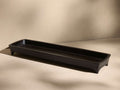 A long, rectangular black tray with slightly raised edges and short, angled legs, positioned on a beige surface.