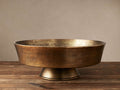 Round, antiqued brass Camden Bowl with pedestal base, made of cast-aluminum, sitting on a rustic wooden table.