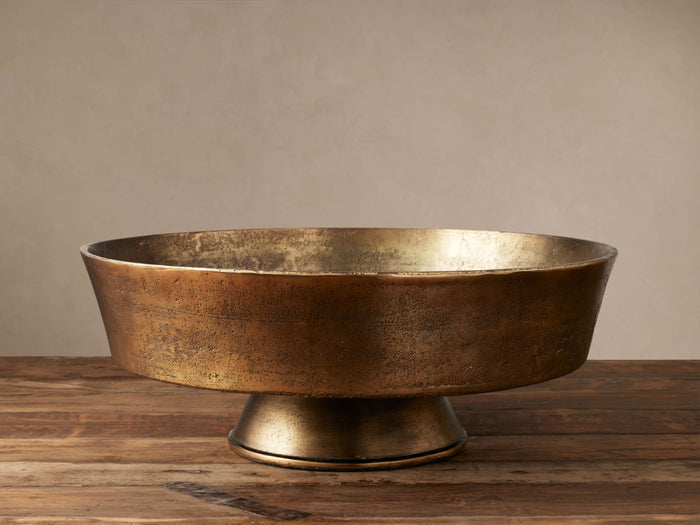 Round, antiqued brass Camden Bowl with pedestal base, made of cast-aluminum, sitting on a rustic wooden table.