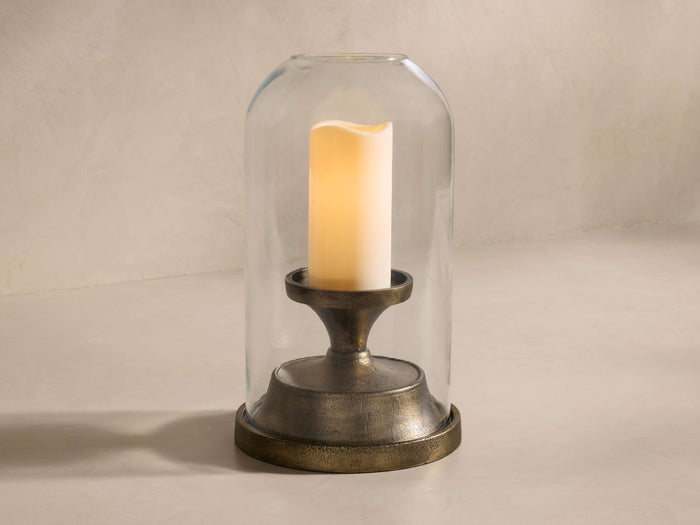 Front view of Camden Hurricane with an antiqued brass base, a clear glass cloche, and a glowing cream pillar candle.