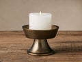 Antiqued brass Camden Pillar Holder with a transitional silhouette, holding a lit white pillar candle on a rustic wooden surface.