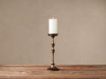 Antiqued brass Camden Tall Pillar Holder, with a transitional silhouette, holding a lit white candle on a rustic wood surface.