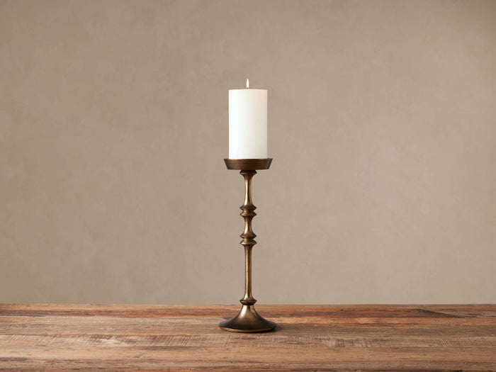 Antiqued brass Camden Tall Pillar Holder, with a transitional silhouette, holding a lit white candle on a rustic wood surface.