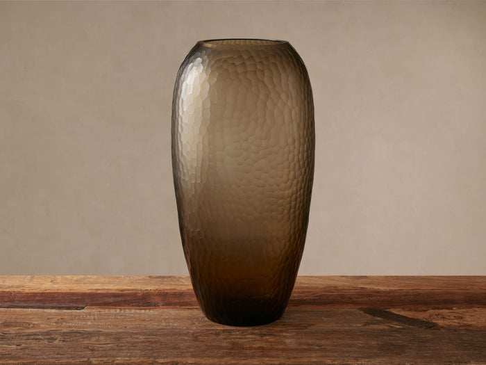 Camille Vase, a tall, smoky grey glass vase with a hammered, rippled texture, stands on a rustic wooden table.
