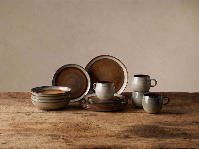 Canyon 16-piece dinnerware set: stacked pasta bowls, plates, and mugs with earthy, reactive brown & neutral glaze on wood.