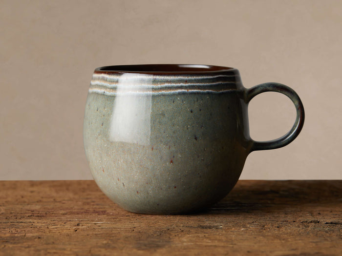 Canyon Mug, hand-glazed stoneware in green-grey with white bands and dark brown rim/handle, on a textured wooden surface.