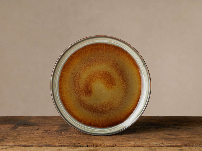 Single Canyon Salad Plate, round, with a reactive amber, gold, brown swirling glaze and a greyish-white rim, on wood.