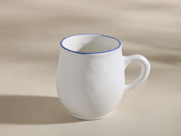 White ceramic Capuchos mug with matte glaze, blue hand-painted rim, and a sturdy handle, angled right on a light surface.