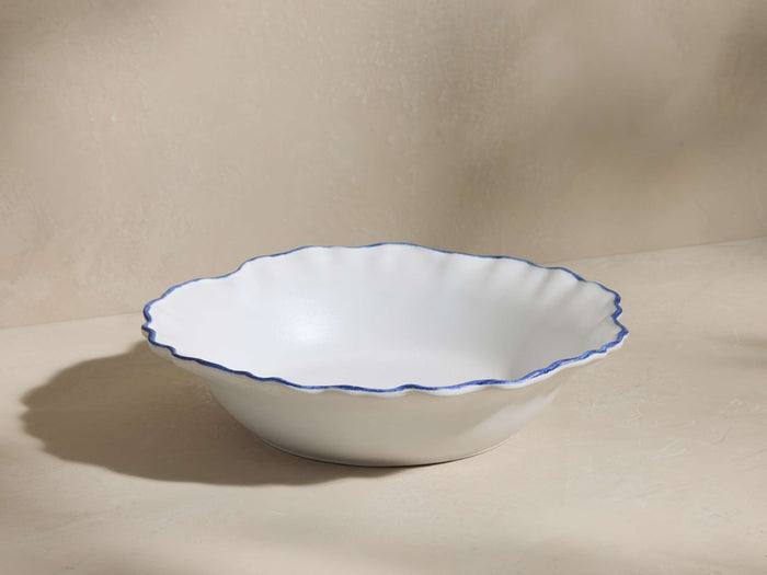 Capuchos Pasta Bowl: White matte ceramic stoneware bowl with a wavy, hand-painted blue rim, angled on a light surface.