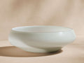 Handcrafted, recycled glass Cassia Bowl, subtle pale green/off-white, organic curved shape, on a light tan background.