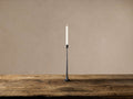 Black cast iron taper candle holder with a sleek shape, standing on a rustic wooden table, holding a lit white candle.