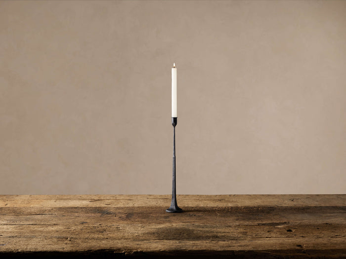 Black cast iron taper candle holder with a sleek shape, standing on a rustic wooden table, holding a lit white candle.