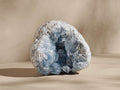 Celestite Mineral: Rough, light blue-grey geode with sparkling deep blue crystals visible inside, on a light beige surface.