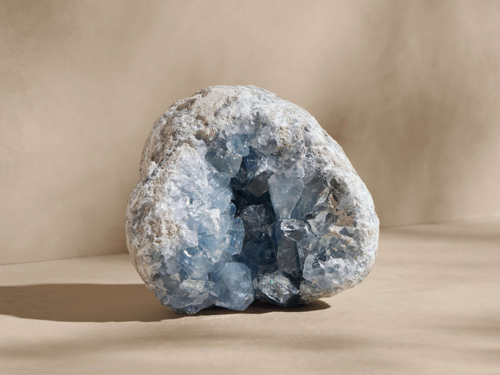 Celestite Mineral: Rough, light blue-grey geode with sparkling deep blue crystals visible inside, on a light beige surface.