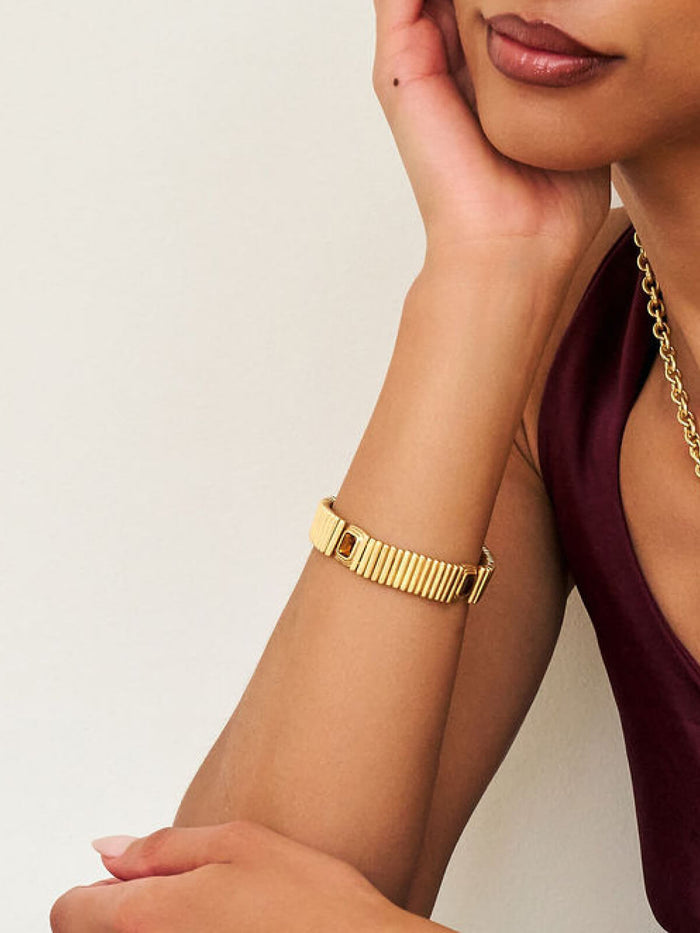A close-up of a woman's arm wearing a gold-toned metal link bracelet, paired with a matching gold chain necklace and a dark burgundy top against a plain off-white background.