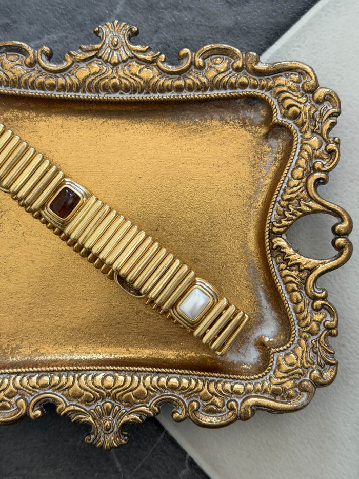 A close-up of a gold-toned ornate tray with a matching ribbed belt featuring small dark and white stones, set against a dark gray and light beige background.