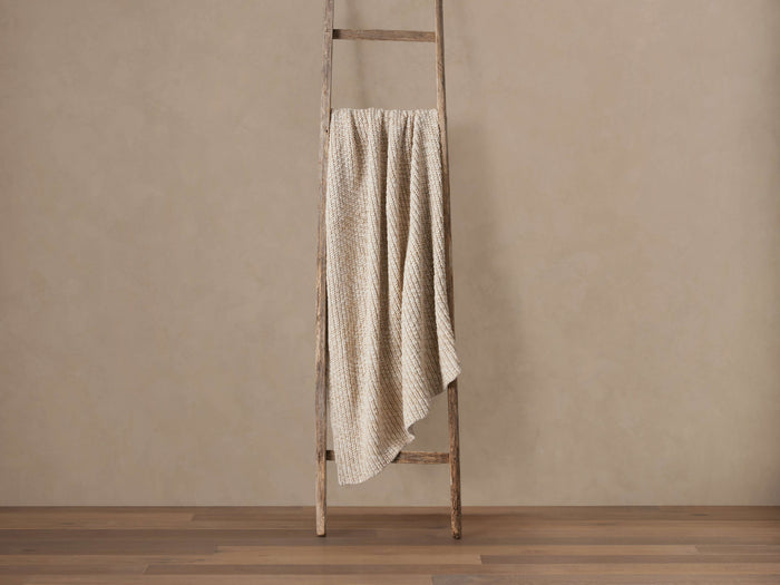 Light beige chunky throw, knitted texture visible, draped vertically over a rustic wooden ladder against a tan wall.